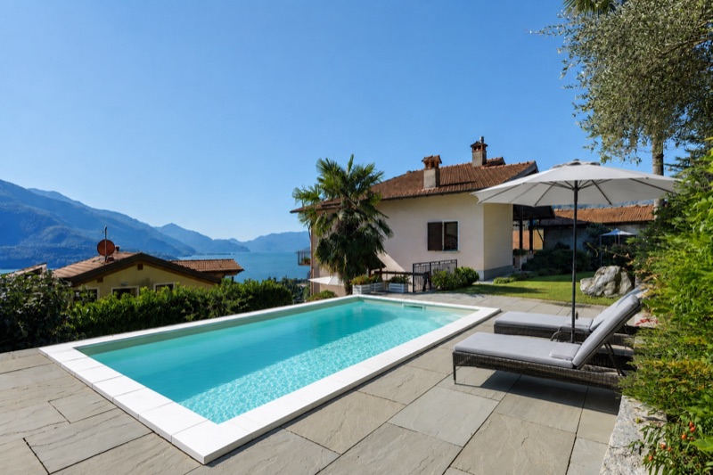 The outdoor pool of Casa Artis with sun loungers, the house behind and Lake Como in the distance