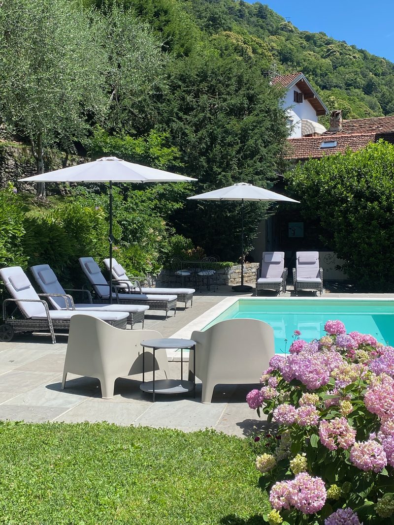 Casa Artis's pool area with loungers and armchairs, hydrangeas in bloom in the foreground and the house on the green hillside behind