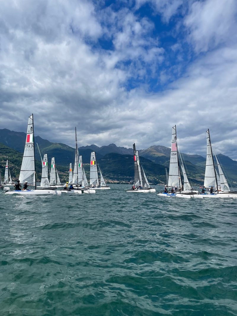 A fleet of small sailing catamarans racing on Lake Como, with team flags and the mountains behind