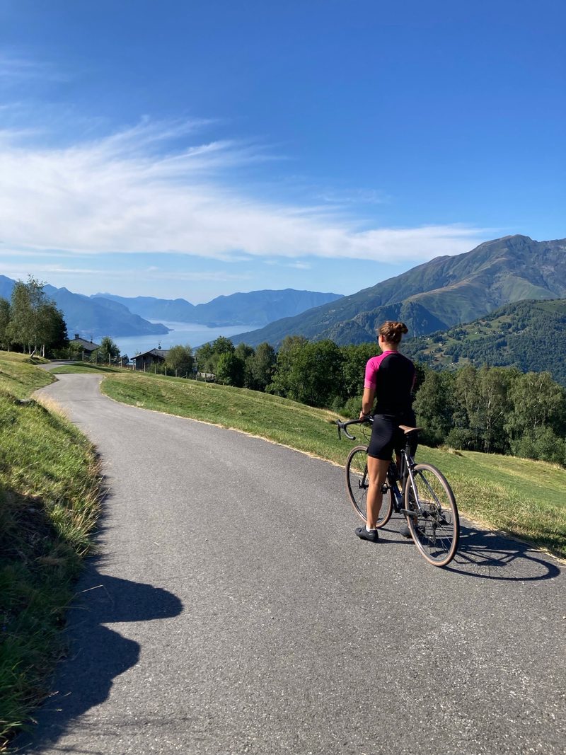 A cyclist on a road bike pausing on a scenic mountain road, with the lake visible far below among the peaks