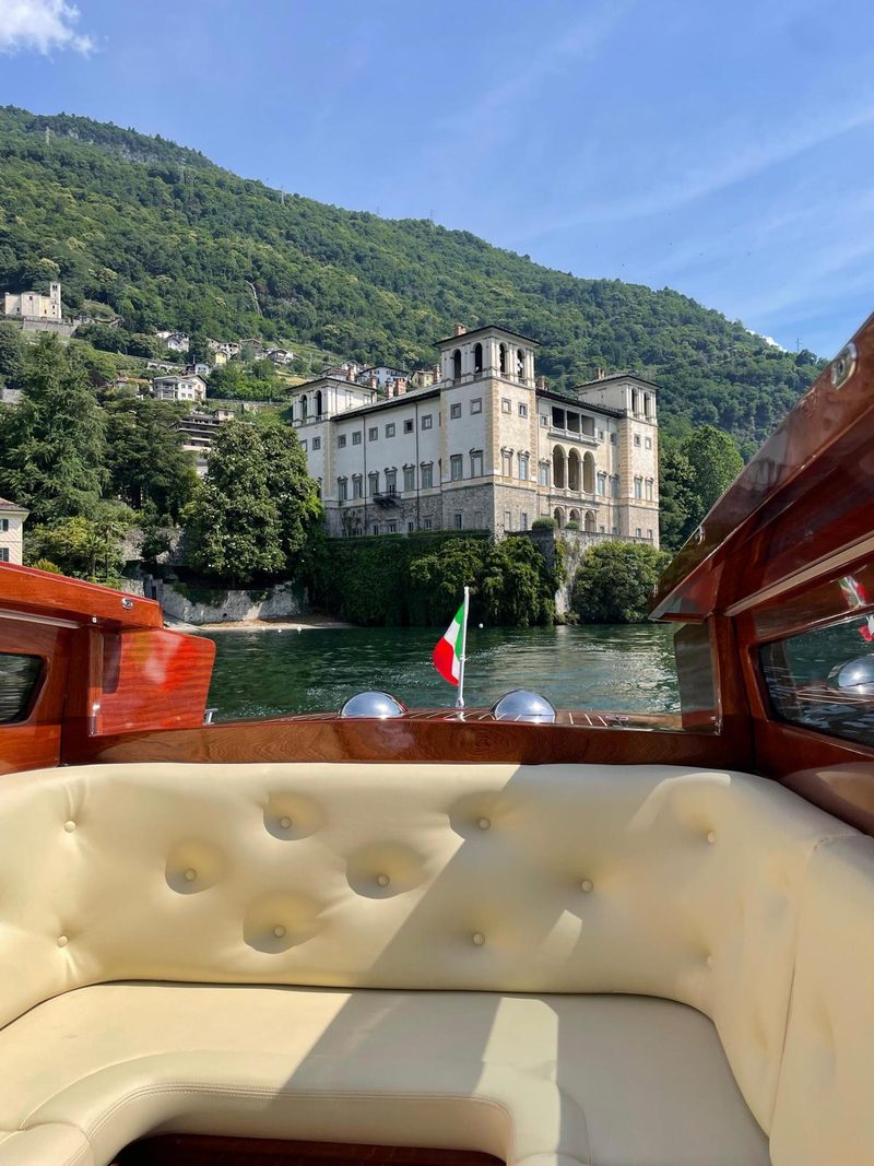 The view from a vintage boat of a historic lakeside palazzo, framed by cypress trees and hills