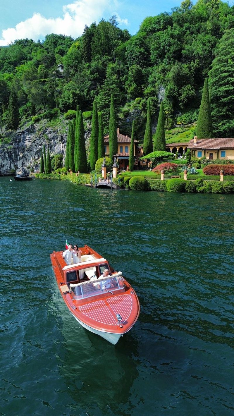 A mahogany motorboat passing a historic villa with a cypress garden on the lakeshore
