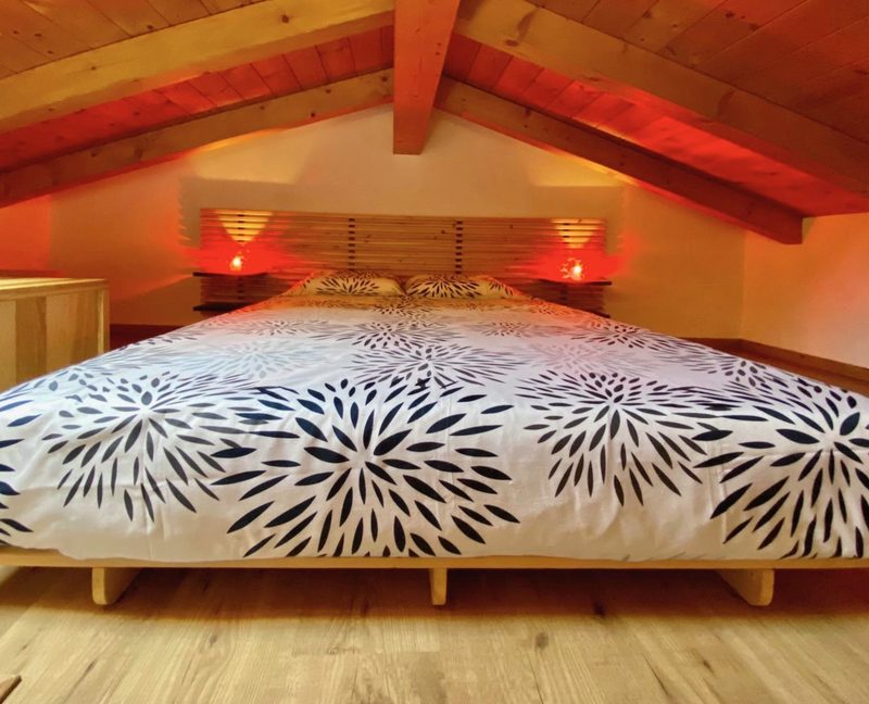 The sleeping loft of Bucaneve under the timber roof, lit by warm bedside lamps