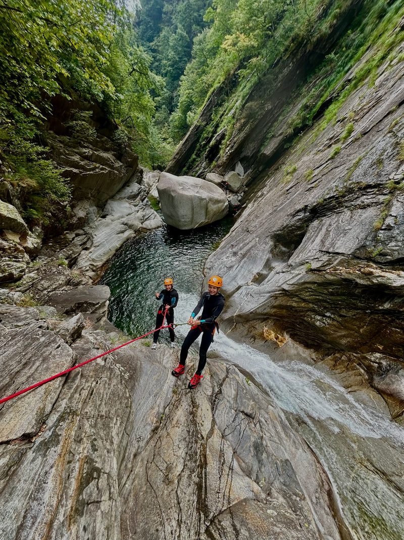 Two people in wetsuits and helmets canyoning, rappelling down the rocks of a mountain stream