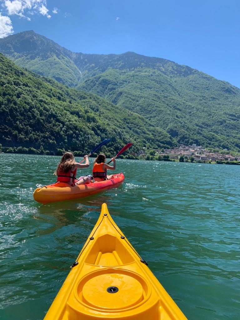 Two people in life jackets paddling a red kayak, seen from another kayak, with mountains and a lakeside village behind