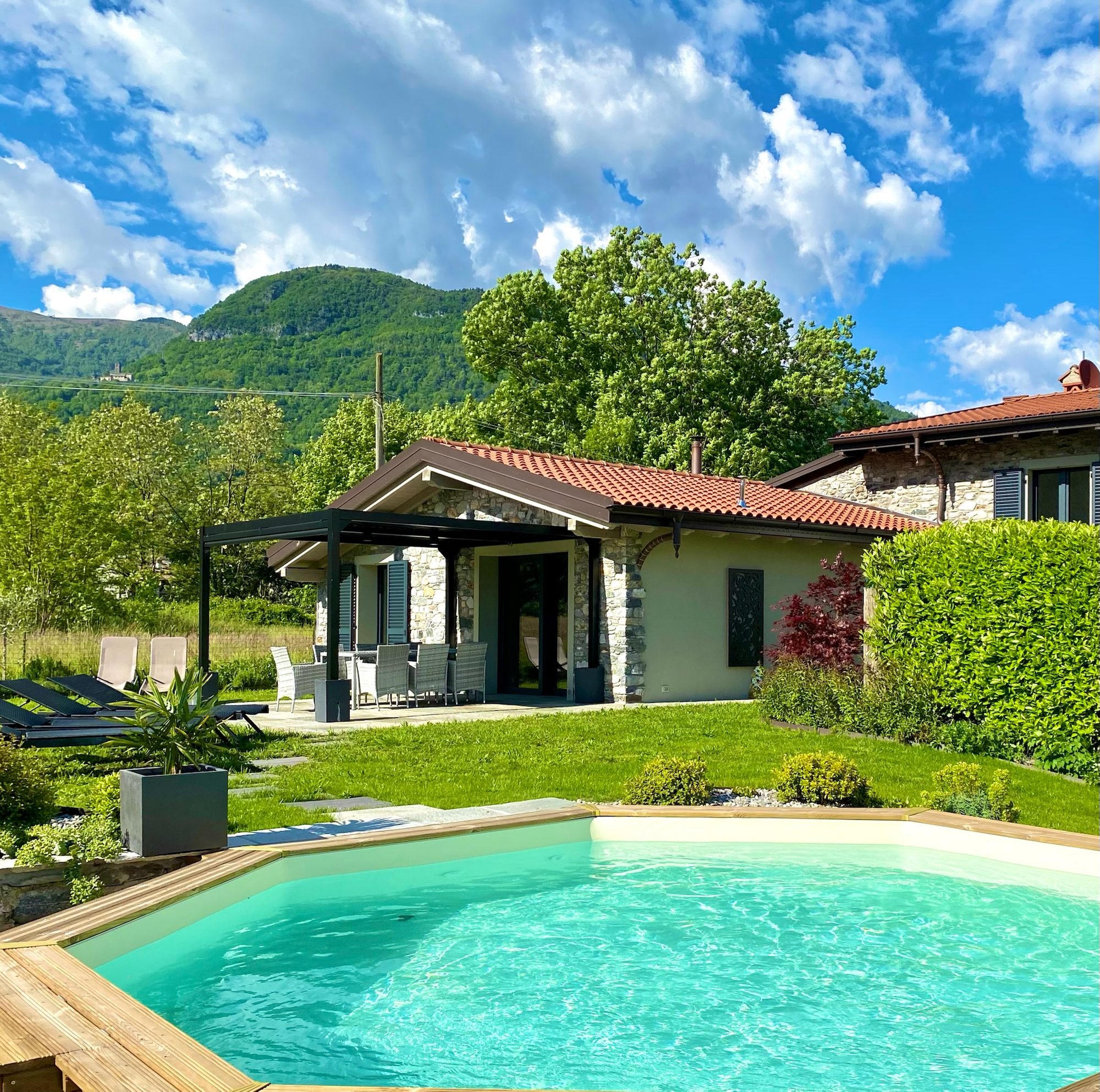Villa Larius under a blue summer sky, its saltwater pool in the foreground and green mountains behind