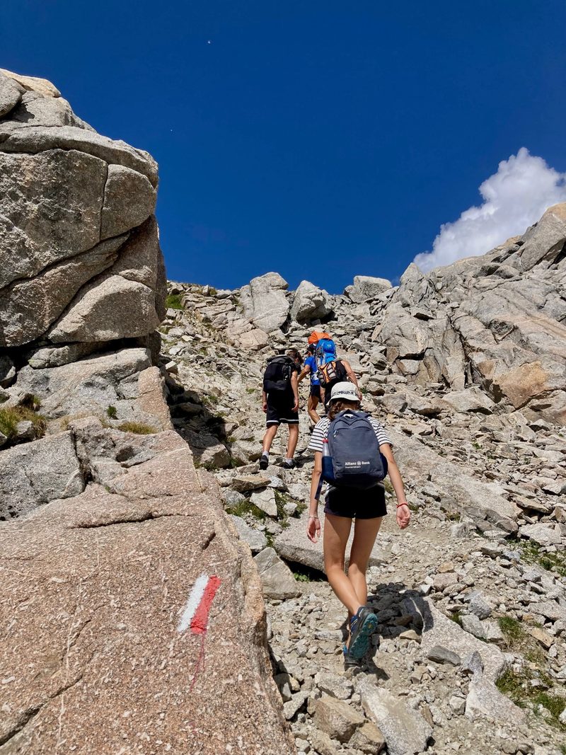 A group of hikers with backpacks climbing a rocky mountain trail under a clear blue sky