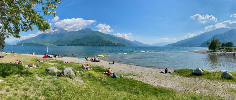 Serenella beach with people relaxing on the grass and pebbles, the lake and the Alto Lario mountains behind