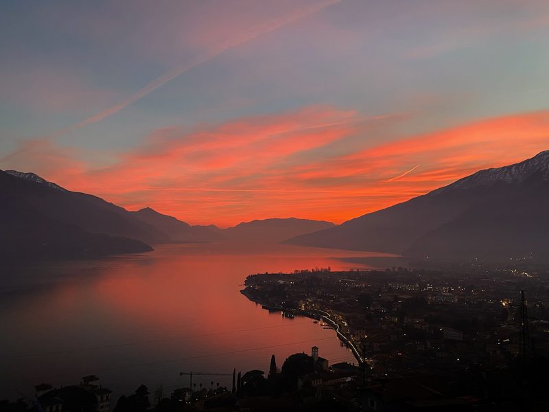 An orange and pink sunset over Lake Como, seen from above Gravedona, with the lit-up lakefront below