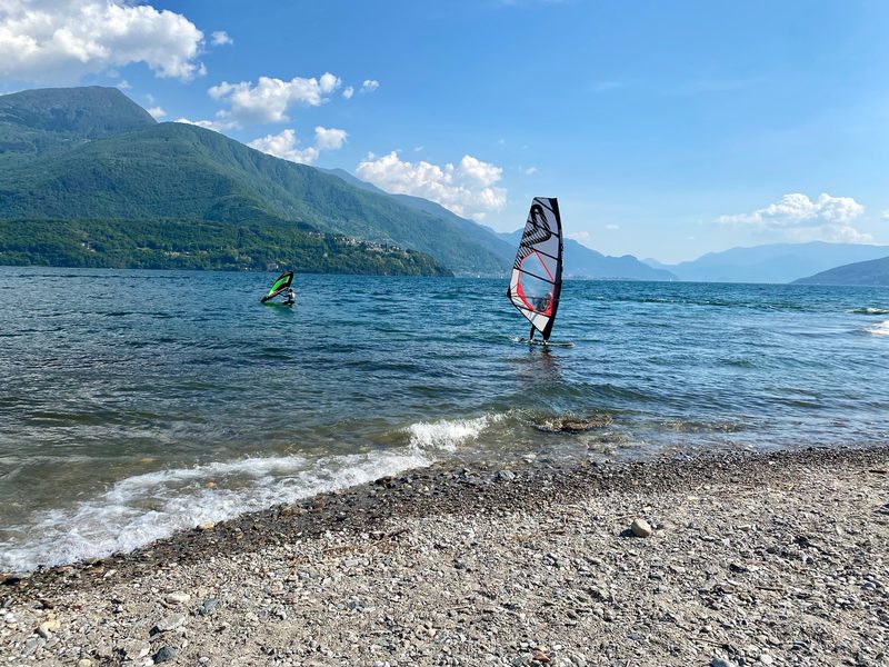 Two windsurfers on the lake seen from a pebble beach, with the Alto Lario mountains behind